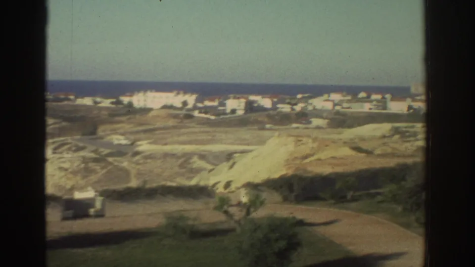 1976 Greece home-movie frame showing a coastal village beyond pale sandy hills, with the sea on the horizon.