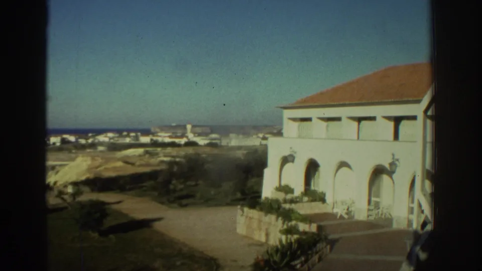 1976 Greece frame with a white arched building in the foreground and a seaside village beyond sandy hills.