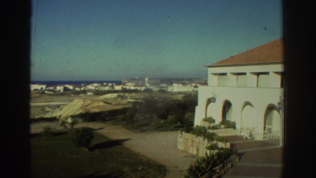1976 Greece 8mm frame showing a white Mediterranean-style house near the coast, with arched windows, a road and distant buildings.
