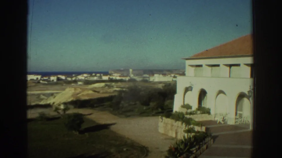 1976 Greece 8mm frame showing a white Mediterranean-style house near the coast, with arched windows, a road and distant buildings.
