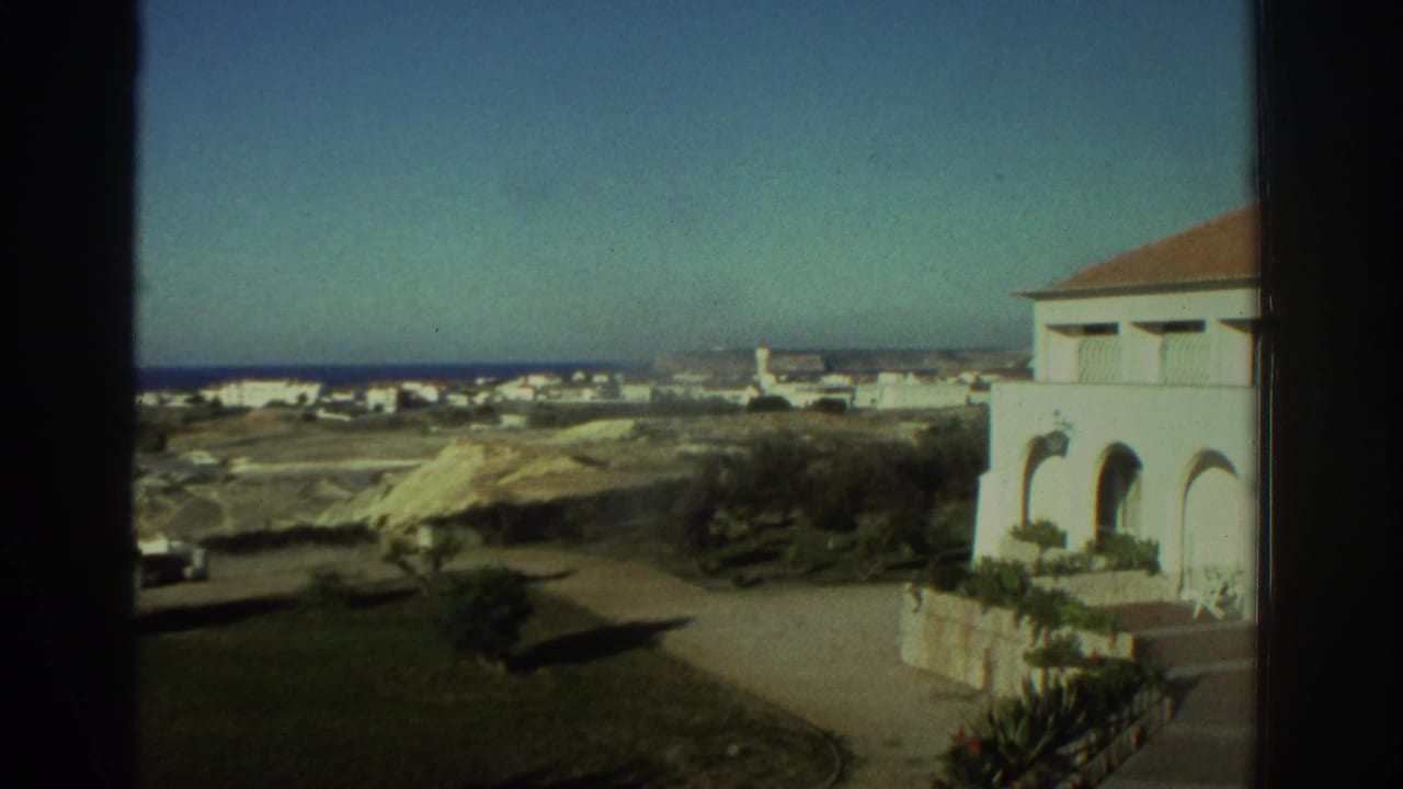1976 Greece home-movie view of a white coastal house, road and low seaside settlement under clear daylight.