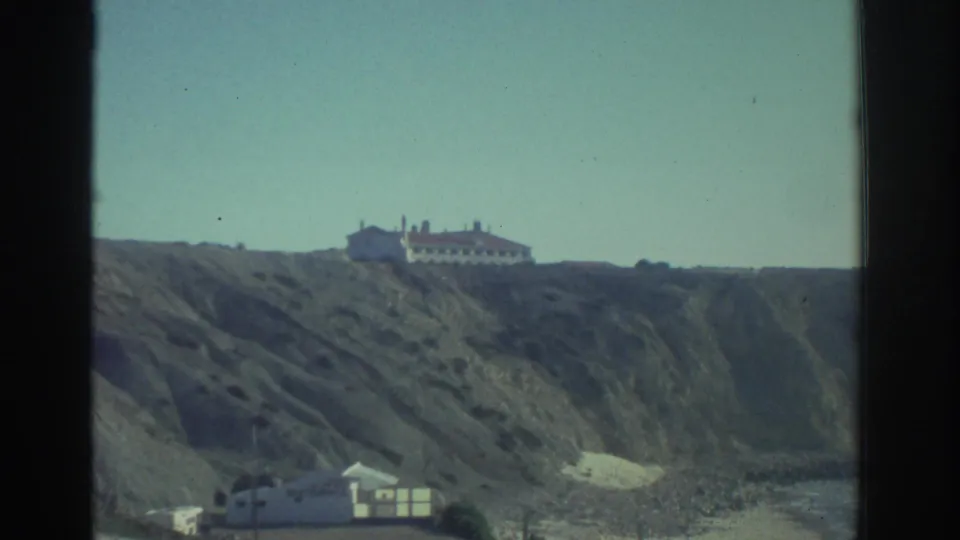 1976 Greece 8mm frame showing a large house on a long cliff, with a lower beach structure and rugged slope below.