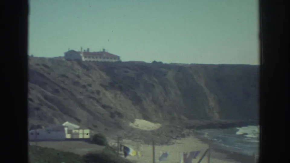 1976 Greece home-movie frame of a cliffside house above a beach area, with shoreline water and small structures below.