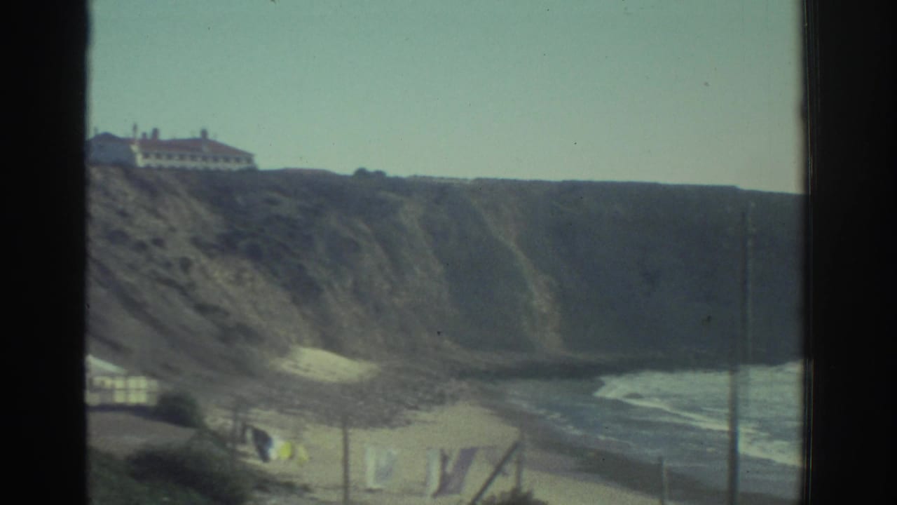 1976 Greece 8mm view through a window frame toward coastal cliffs, a sandy beach, laundry near shore, and a white building above the slope.