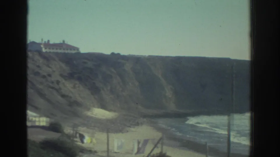 1976 Greece 8mm view through a window frame toward coastal cliffs, a sandy beach, laundry near shore, and a white building above the slope.