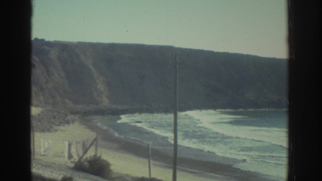 1976 Greece travel footage frames rugged coastal cliffs, a sandy beach, rolling surf, and a vertical pole through a dark window mullion.