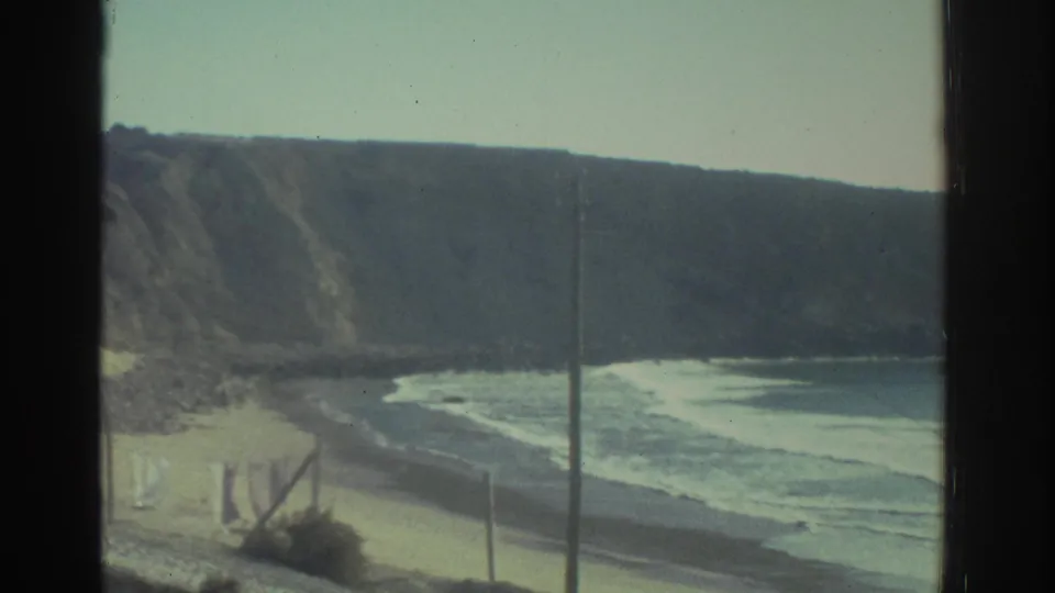 1976 Greece travel footage frames rugged coastal cliffs, a sandy beach, rolling surf, and a vertical pole through a dark window mullion.