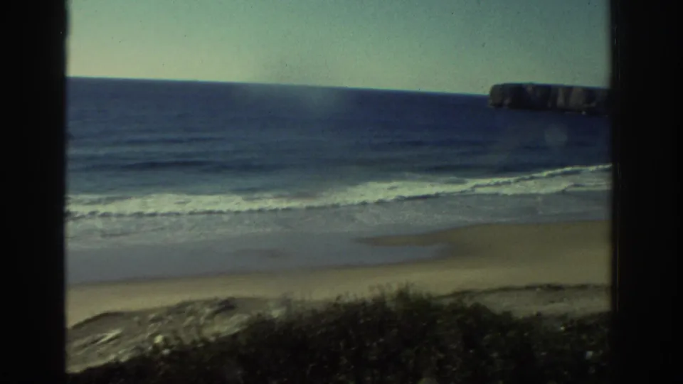 1976 Greece 8mm frame showing a sandy beach, gentle surf, blue sea, low foreground brush, and a distant rocky headland through a window.
