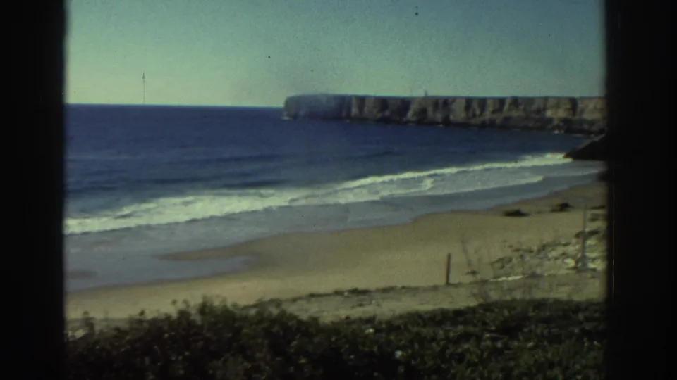 1976 Greece coastal view through window glass, with empty sand, soft surf, blue water, foreground vegetation, and a cliff-like headland.
