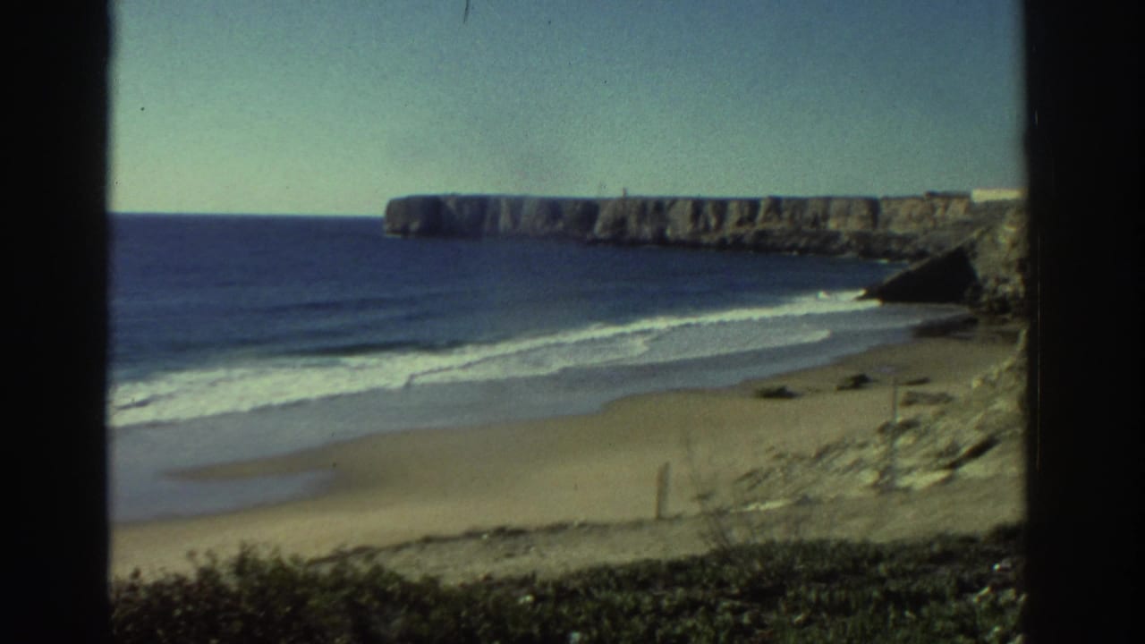 1976 Greece 8mm frame showing surf rolling onto a sandy beach beneath a broad rocky coastal cliff.