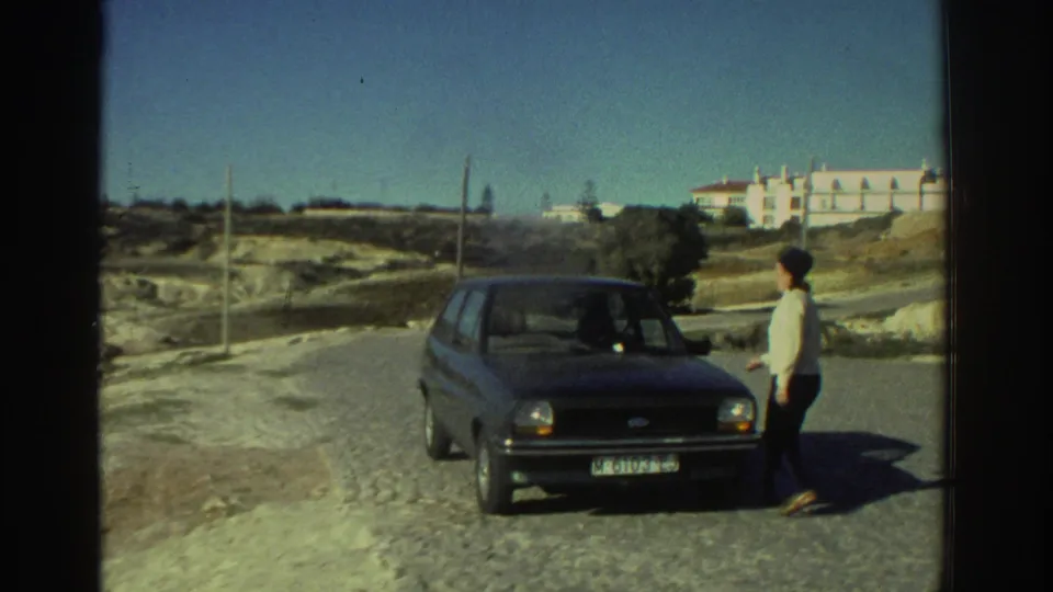 1976 Greece frame with a man beside a small dark hatchback on a gravel roadside, with buildings in the background.