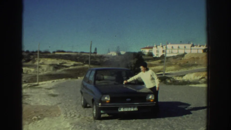 1976 Greece home-movie frame showing a man leaning toward the front of a small hatchback on a gravel road.