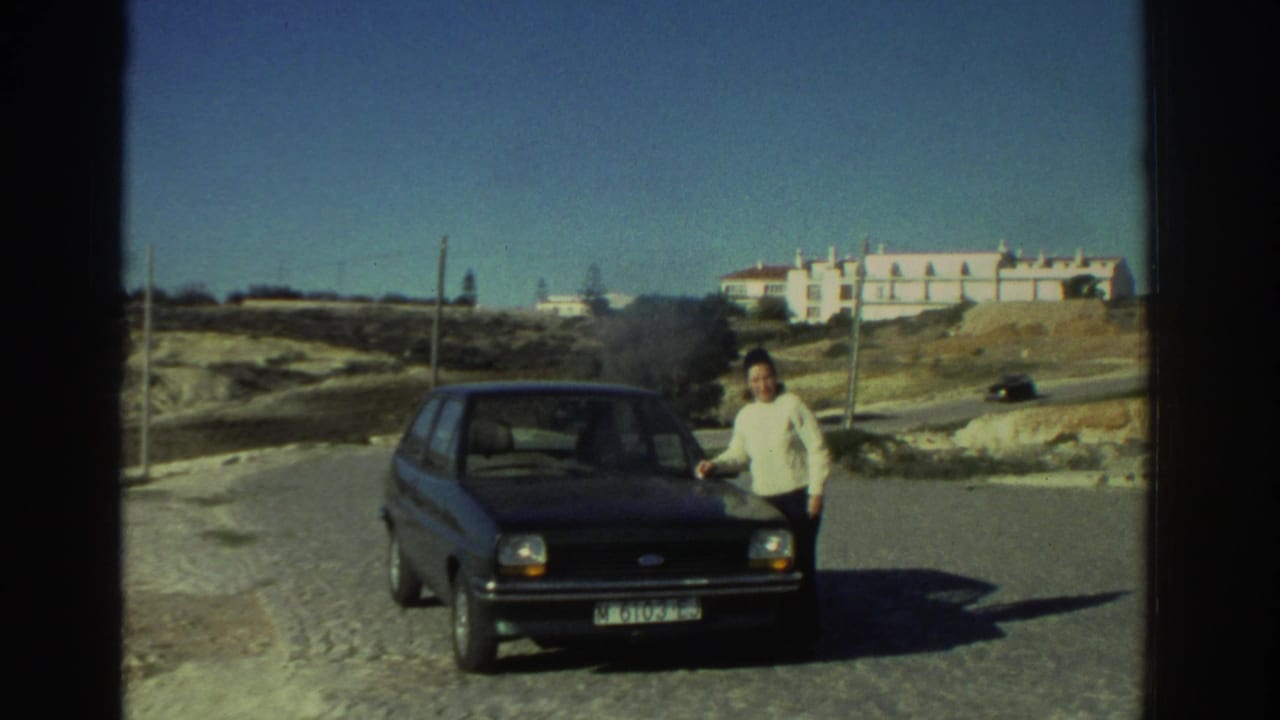 1976 Greece home-movie frame showing a woman beside a dark compact car on a cobblestone road near arid hills and a pale building.