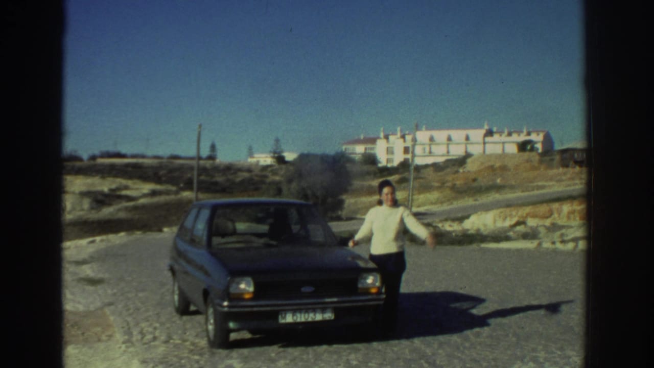 Woman gesturing beside a dark compact car on a road with a pale building and dry hills beyond.