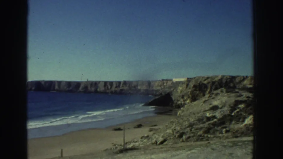 1976 Greece home-movie frame looking over a sandy beach, deep blue sea, rocky foreground, and pale cliffs under clear sunlight.
