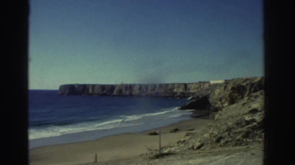 1976 Greece travel frame showing a quiet beach below cliffs, with blue water, rocky slopes, and bright open sky.