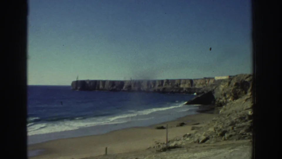 1976 Greece 8mm frame showing a sandy beach, blue sea, pale sky, and rocky cliffs viewed through dark vertical frame edges.