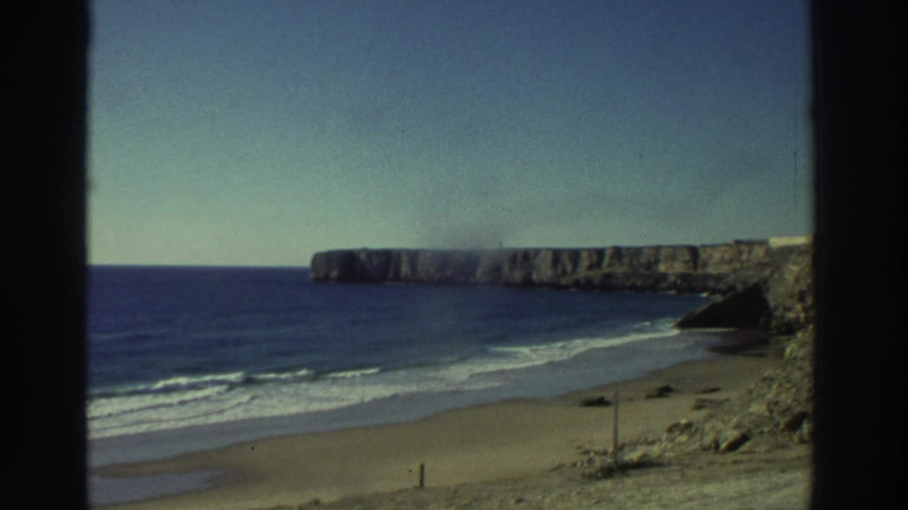 1976 Greece 8mm frame with surf lines on a sandy beach, rocky cliff faces, open water, and a clear pale sky.