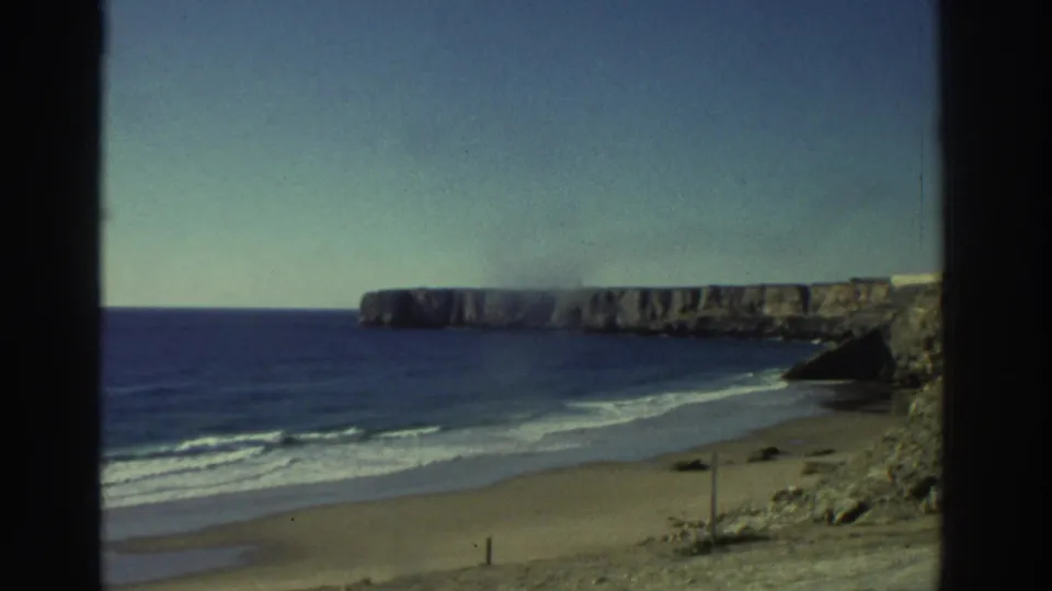 1976 Greece 8mm frame with surf lines on a sandy beach, rocky cliff faces, open water, and a clear pale sky.
