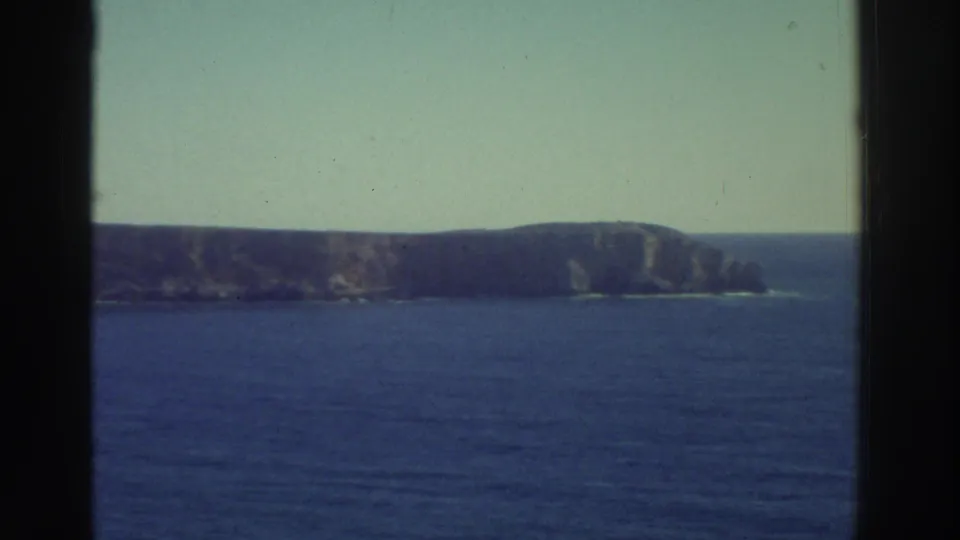 1976 Greece 8mm frame showing open blue water and a distant cliff coastline, with soft grain and faded color.
