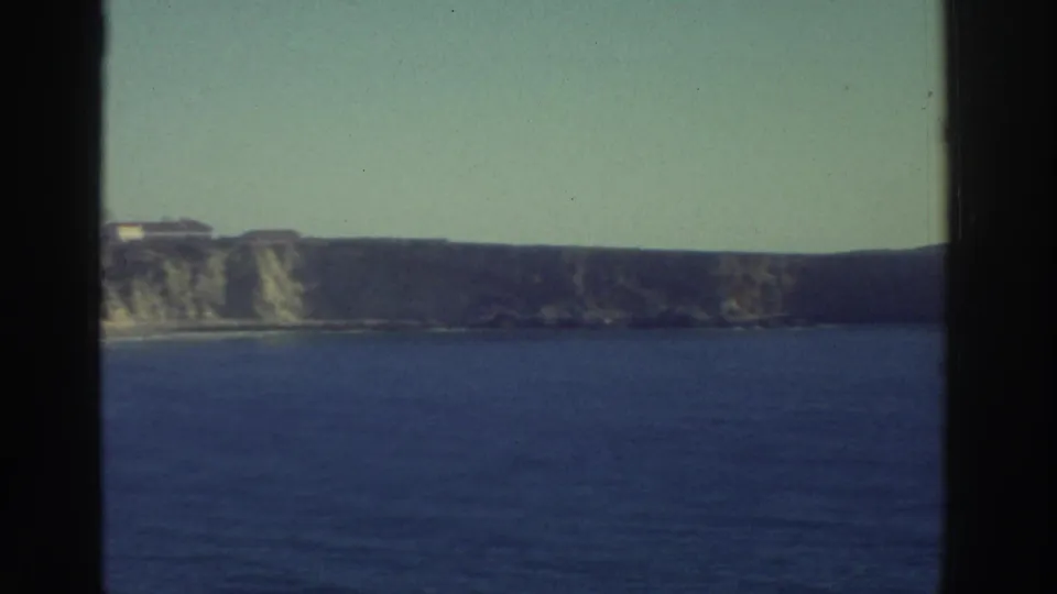 1976 Greece 8mm frame of a rocky coastline across the water, with small distant cliff-top structures and pale sky.
