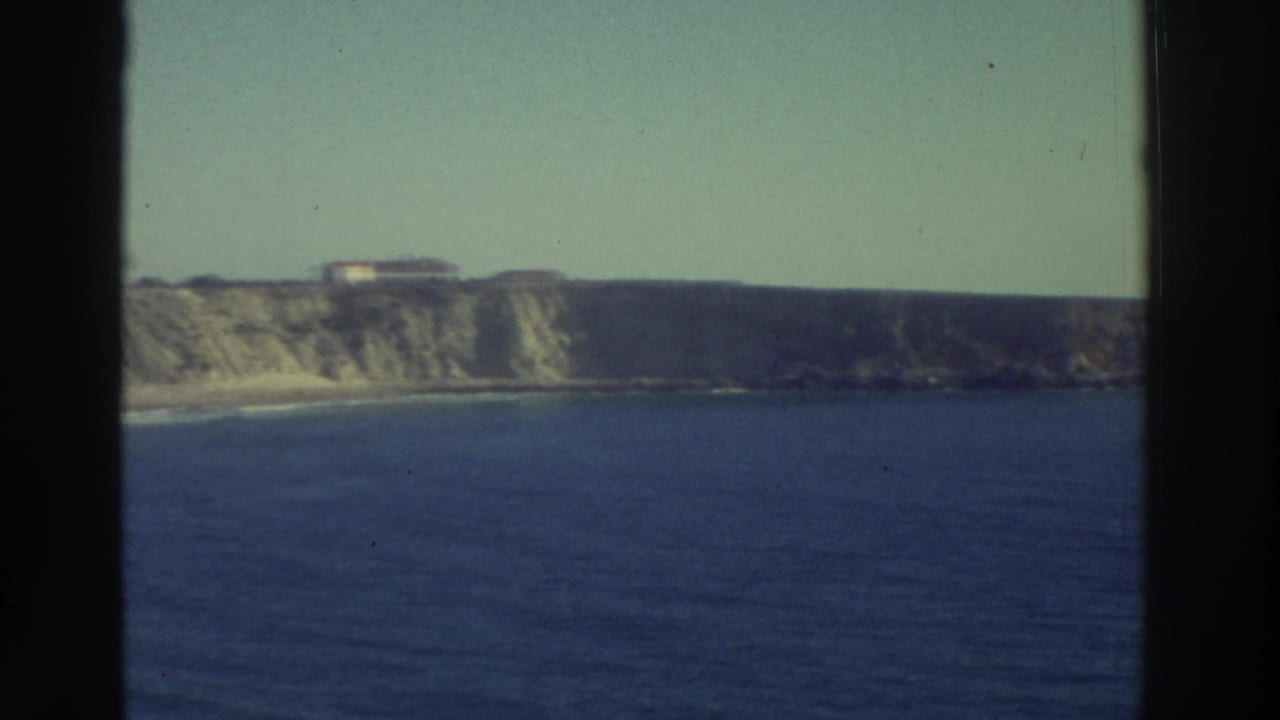 1976 Greece 8mm frame showing pale coastal cliffs, deep blue water, and a distant building along the ridge.