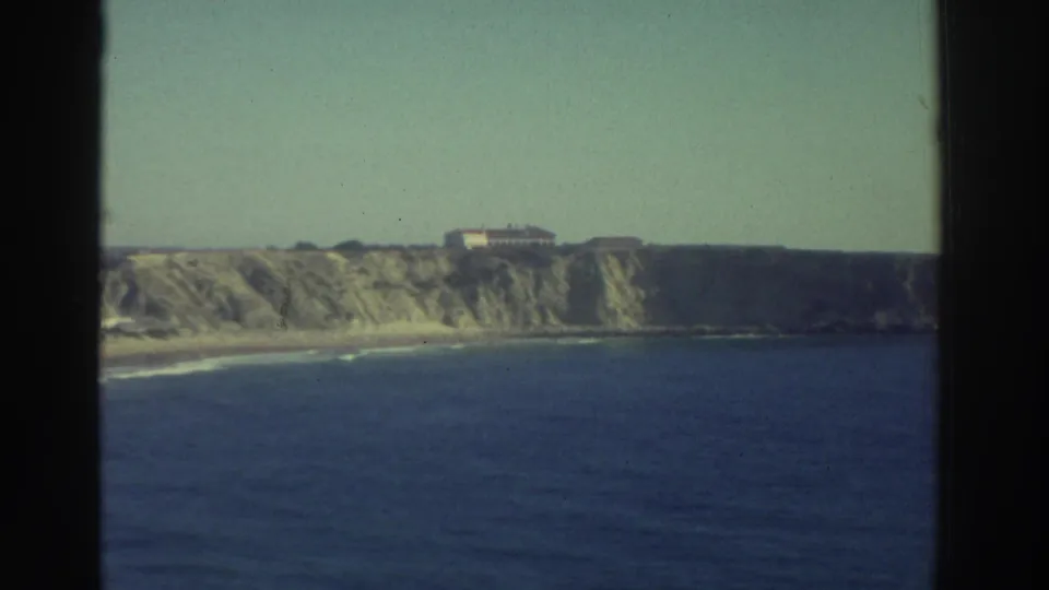 1976 Greece 8mm frame with a broad sea foreground, pale cliffs, light surf, and a small structure on the ridge.