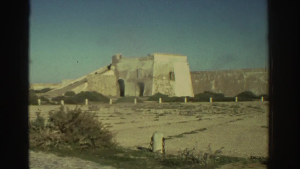 1976 Greece 8mm frame showing a pale stone fortress or castle wall with arches, scrub, dry ground, and blue sky.
