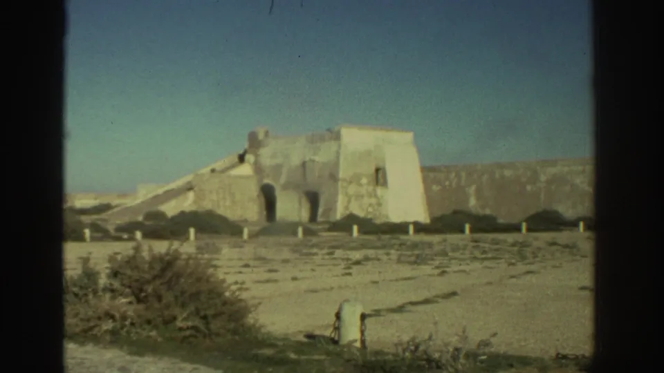 1976 Greece 8mm frame of a pale fortress wall with arched openings, low vegetation, posts, and open sky.