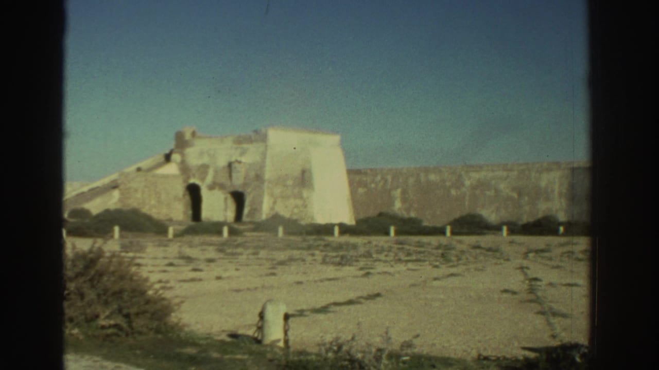 1976 Greece 8mm frame showing a fortress-like stone wall, pale masonry, sandy ground, sparse shrubs, and dark film edges.