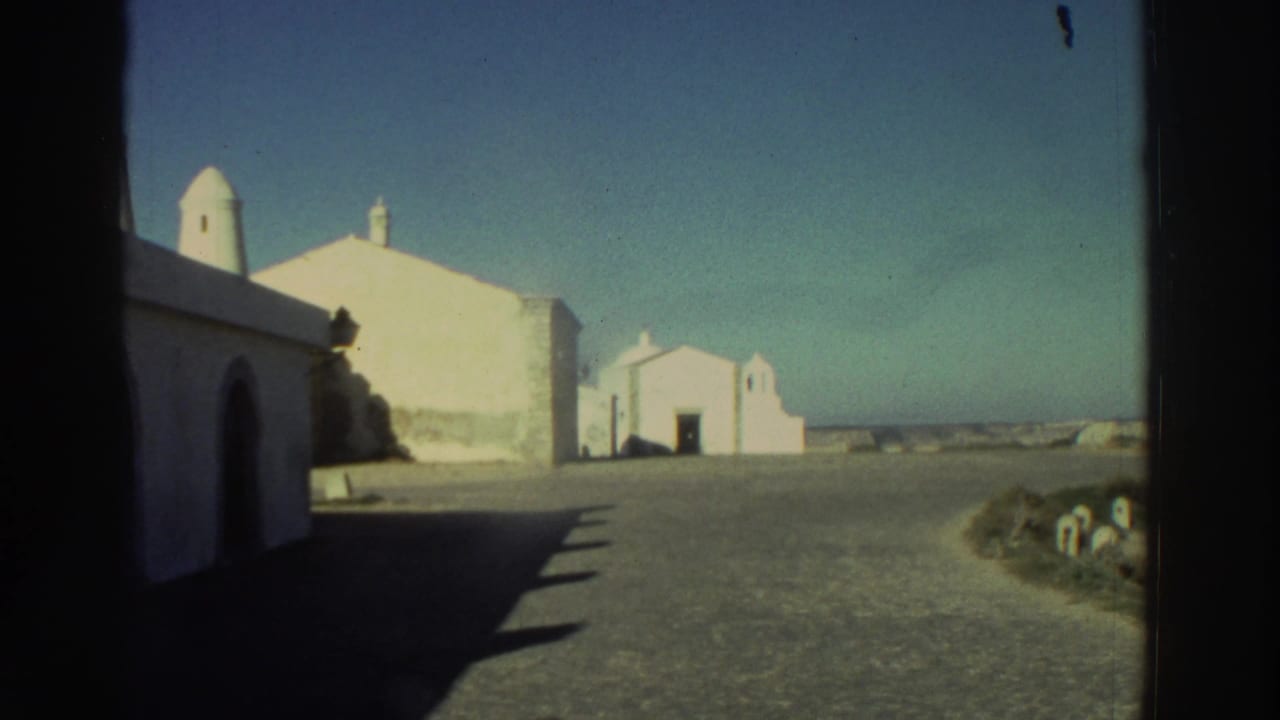 1976 Greece 8mm frame with whitewashed buildings, arched doorway, small chapel-like structure, open ground, and sea or horizon beyond.