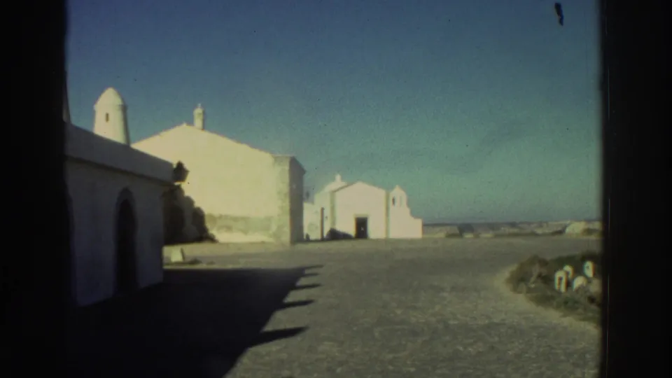 1976 Greece 8mm frame with whitewashed buildings, arched doorway, small chapel-like structure, open ground, and sea or horizon beyond.