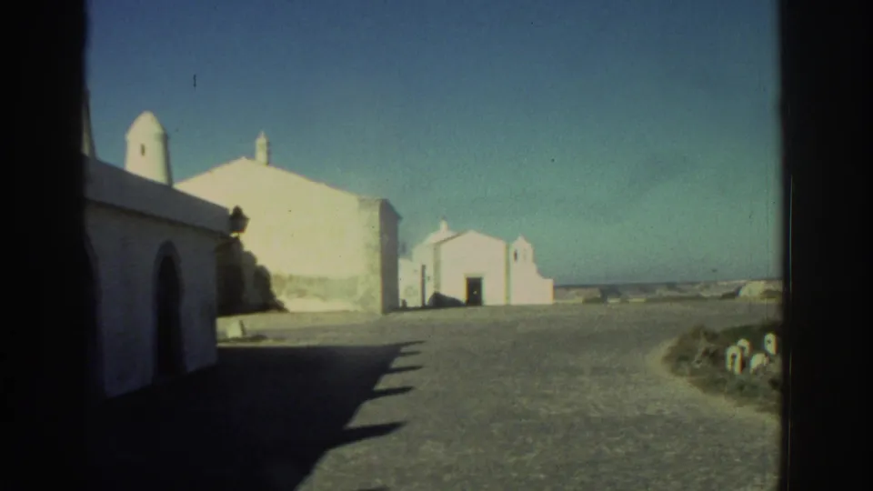 1976 Greece 8mm frame showing whitewashed structures, a shadowed arched opening, small chapel-like building, and sparse roadside vegetation.