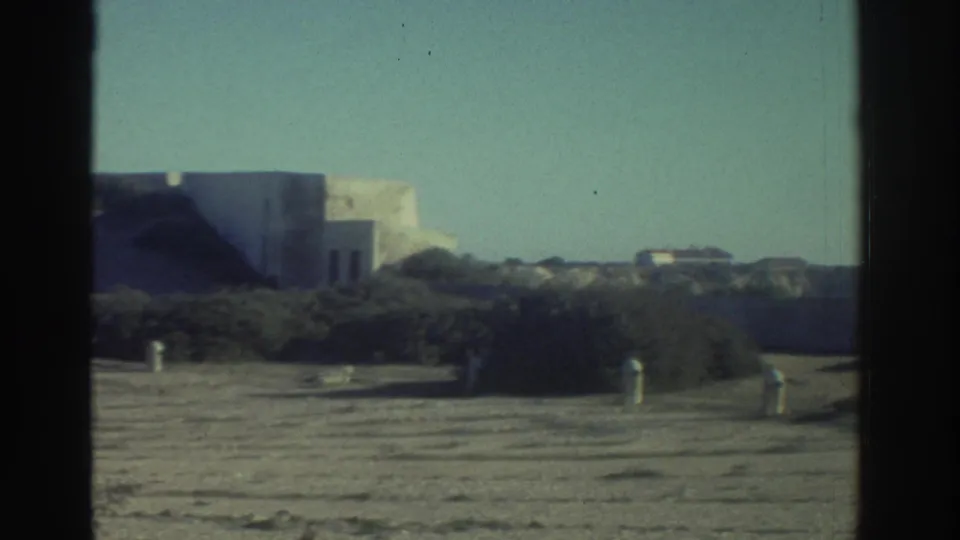 1976 Greece 8mm frame with low pale buildings, scrub vegetation, sandy ground, and a dark vertical film or window edge.