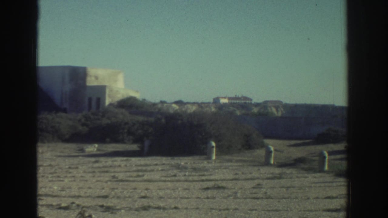 1976 Greece home-movie frame showing a rocky coastal approach with low buildings and dark 8mm frame borders.