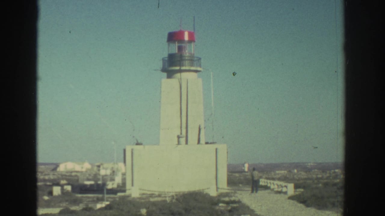 A white lighthouse with a red lantern top stands near the rocky Greek coast under a clear sky.