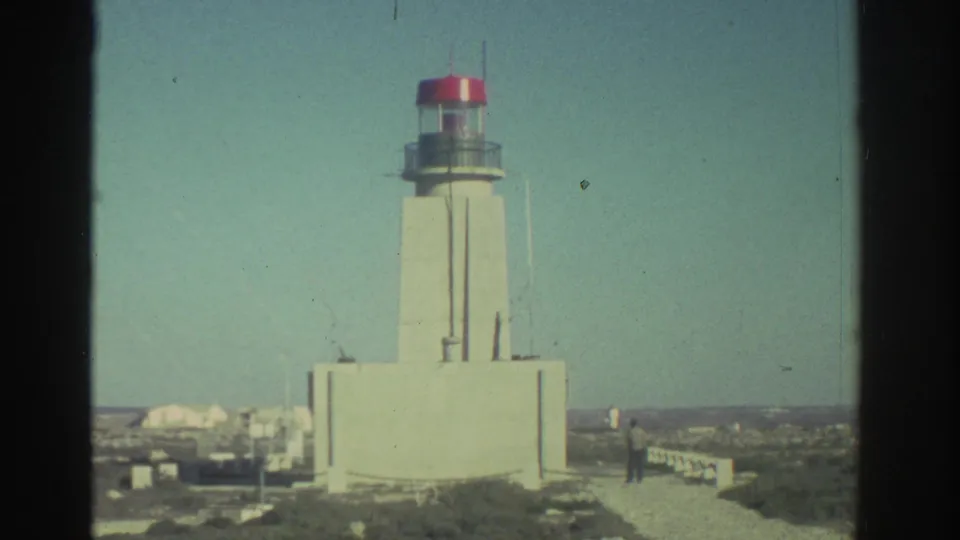 A white lighthouse with a red lantern top stands near the rocky Greek coast under a clear sky.
