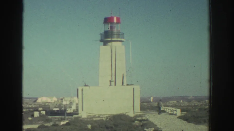 The coastal lighthouse fills the frame, with 8mm grain, faded color, and a distant visitor near the path.