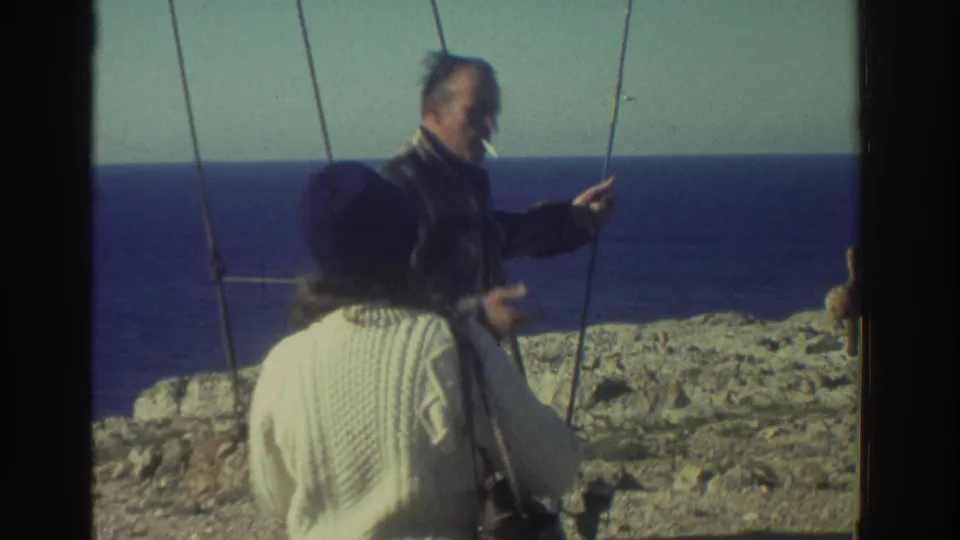In Greece in 1976, a woman in a white sweater and dark hat faces a man near the blue sea on a windy coast.