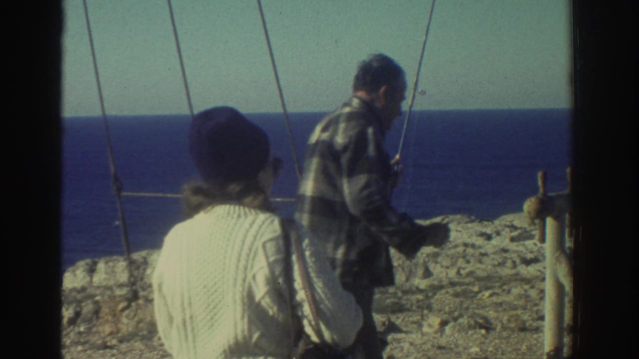 1976 Greece home-movie frame showing a man with a fishing rod near a woman in a white sweater on a rocky coast above blue water.