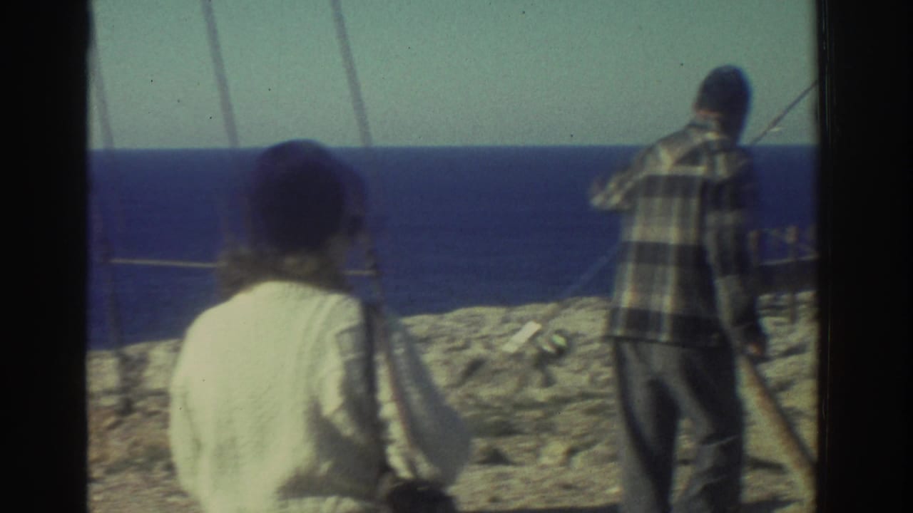 1976 Greece coastal home-movie frame with a woman in a dark hat and a man handling a fishing rod near the blue sea.