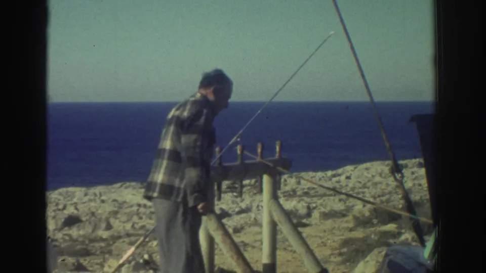 1976 Greece 8mm frame showing a man in a plaid shirt tending a fishing rod near a simple wooden rack on a seaside cliff.