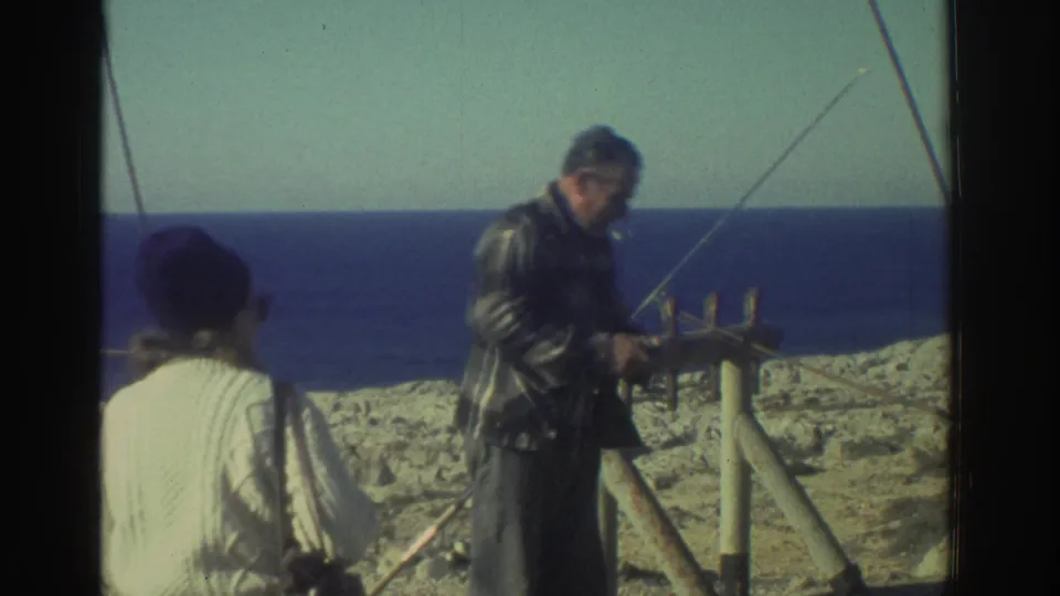 1976 Greece home-movie frame of a man adjusting fishing gear beside a woman on a rocky coastal overlook.