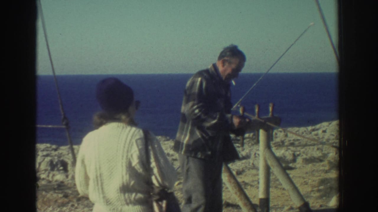 In Greece in 1976, two adults stand on a pale rocky shoreline while one works near a pole-and-frame setup facing the sea.