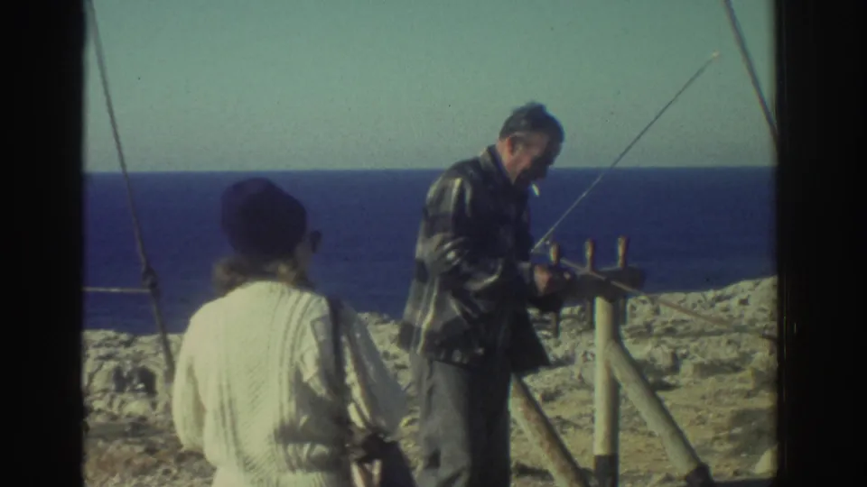 In Greece in 1976, two adults stand on a pale rocky shoreline while one works near a pole-and-frame setup facing the sea.