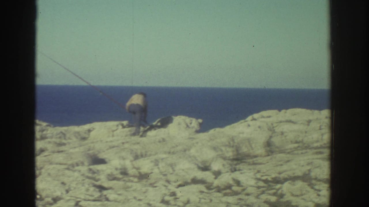 A 1976 Greece shoreline view shows pale rocks, deep blue sea, and a small distant figure or object near the coast within visible film borders.