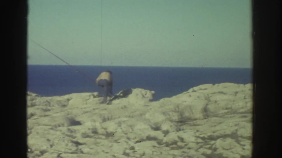A 1976 Greece shoreline view shows pale rocks, deep blue sea, and a small distant figure or object near the coast within visible film borders.