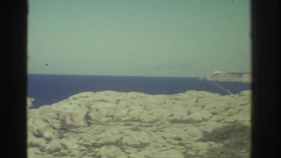 A soft 8mm view of the Greece shoreline shows pale rocks, blue sea, and a long horizontal apparatus or structure partly visible near the horizon.