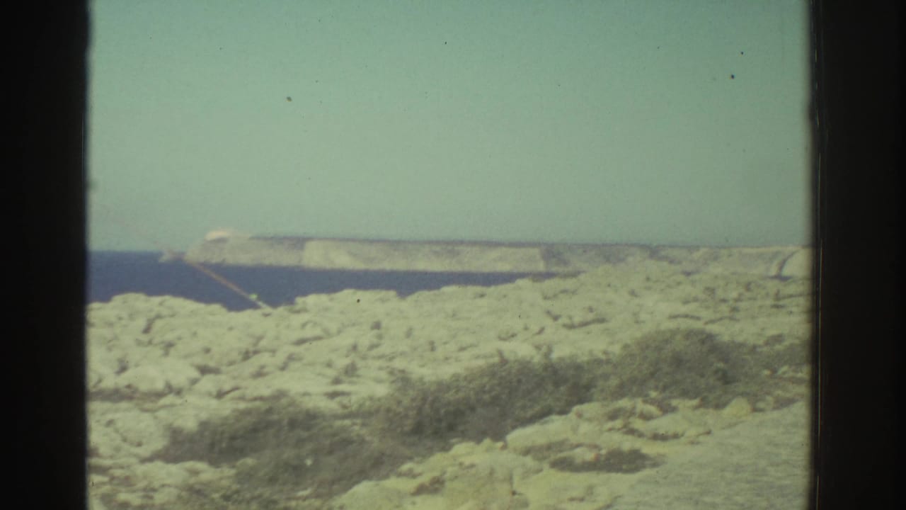 1976 8mm Greece footage showing a rocky coastal edge, deep blue water, and a distant pale headland under bright light.