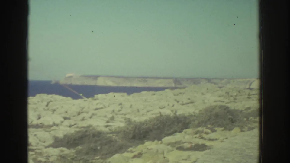 1976 8mm Greece footage showing a rocky coastal edge, deep blue water, and a distant pale headland under bright light.